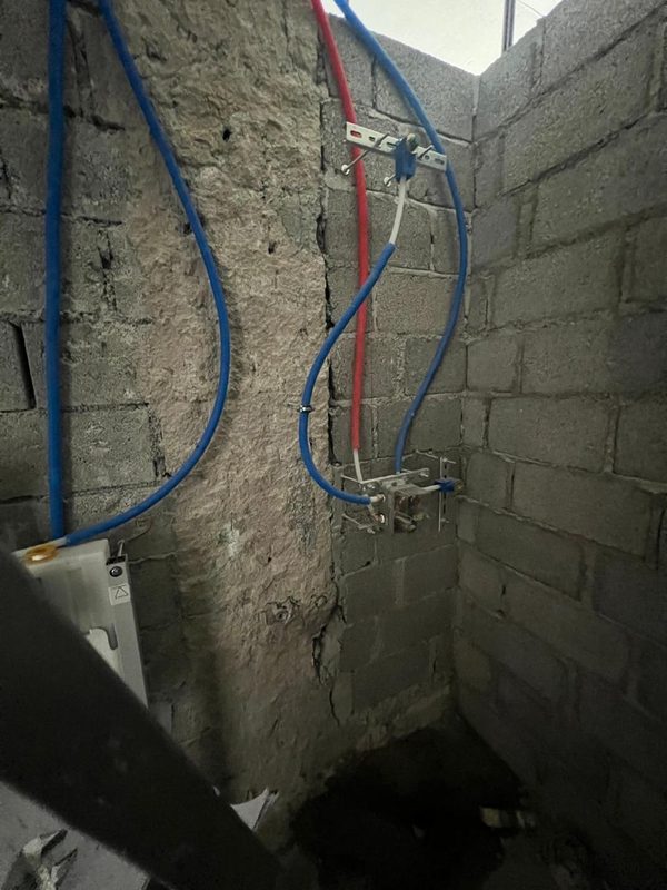Master bathroom during the works: blue and red supply pipes running down a blockwork wall to a valve