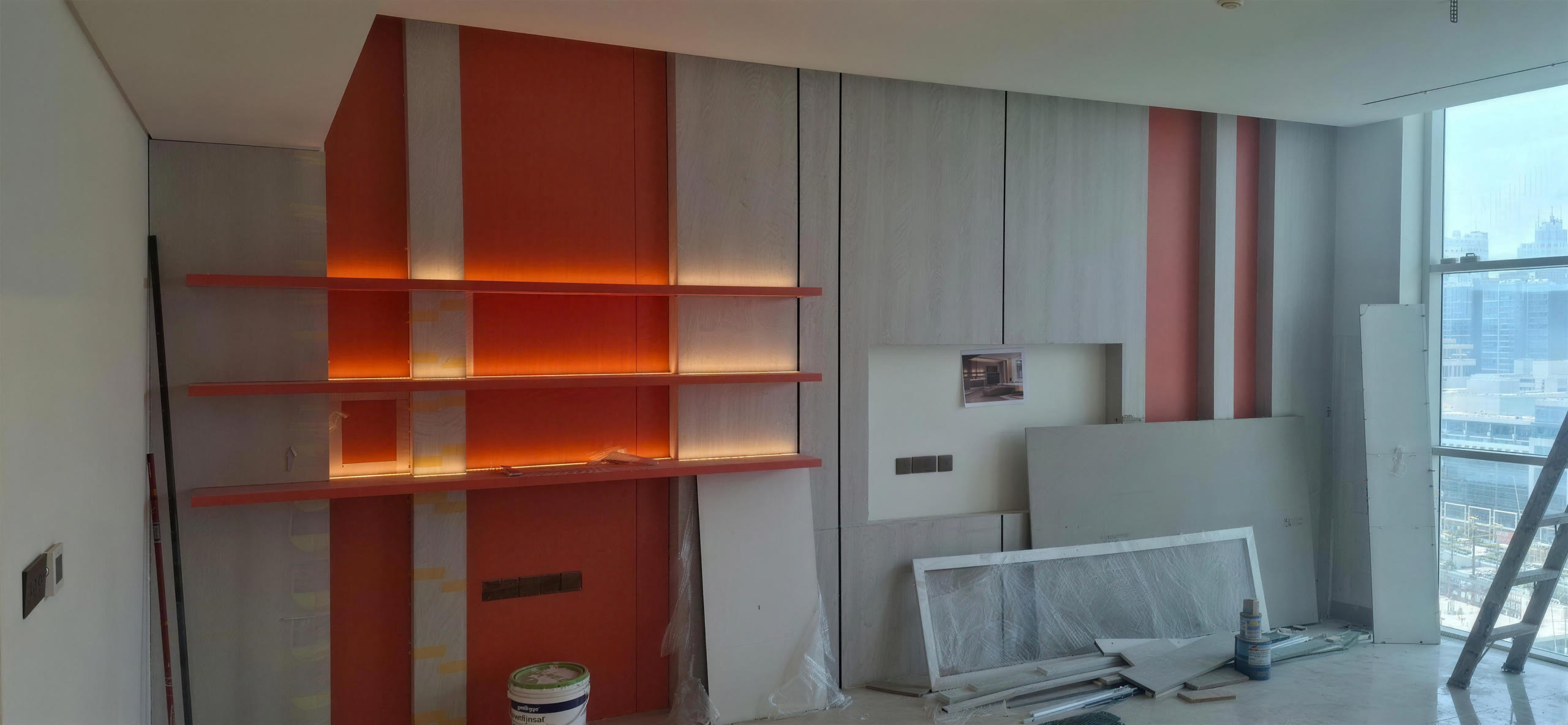 TV feature wall during installation: terracotta lacquer and grey wood panels, backlit shelves and full-height glazing on the right